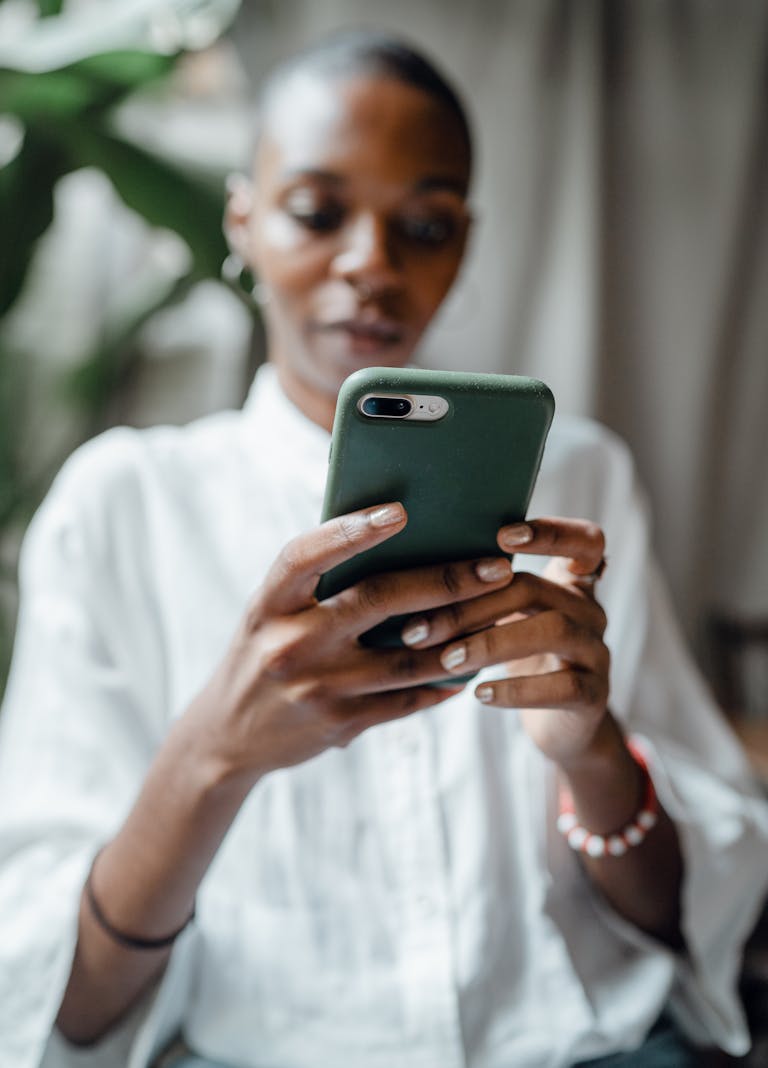Blurred concentrated bald black female in white casual clothes using smartphone in green cover while sitting near big plant in daytime