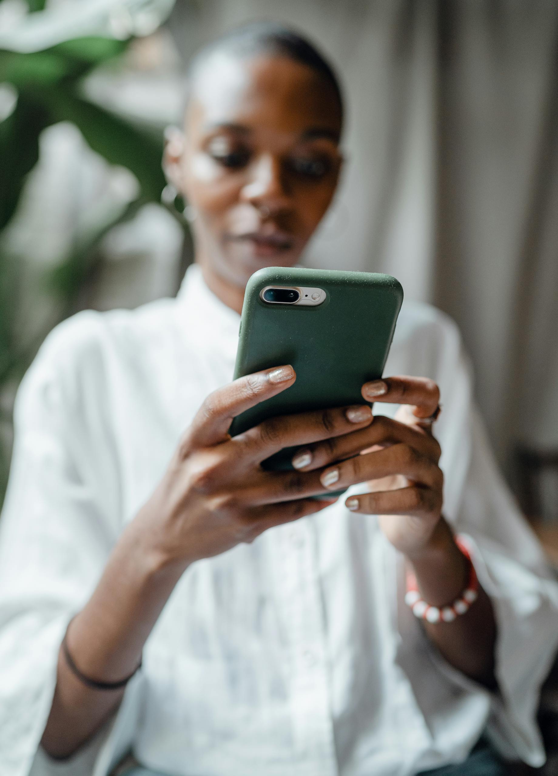 Blurred concentrated bald black female in white casual clothes using smartphone in green cover while sitting near big plant in daytime